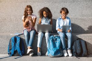 Smiling school kids sitting with digital devices at the street